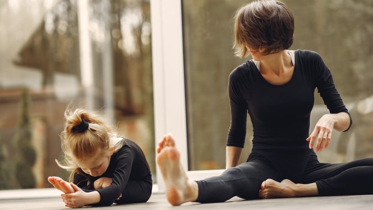 Anonymous female instructor in sportswear sitting near little blond gymnast doing tilt forward on floor near transparent door while stretching legs